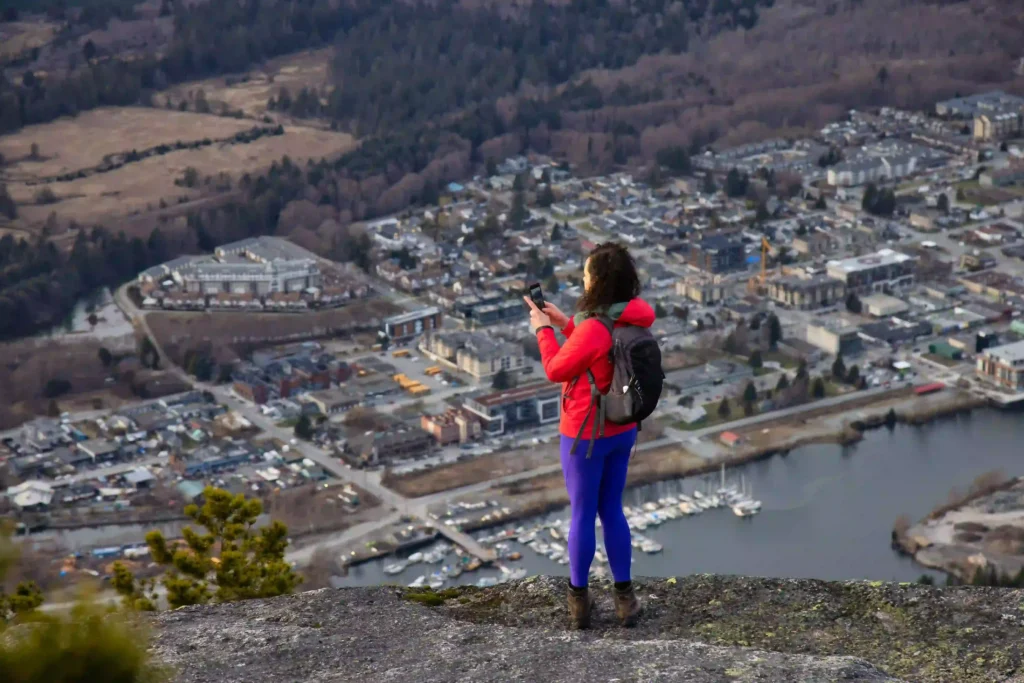 A woman in a red jacket captures a city and mountain view from a scenic overlook.